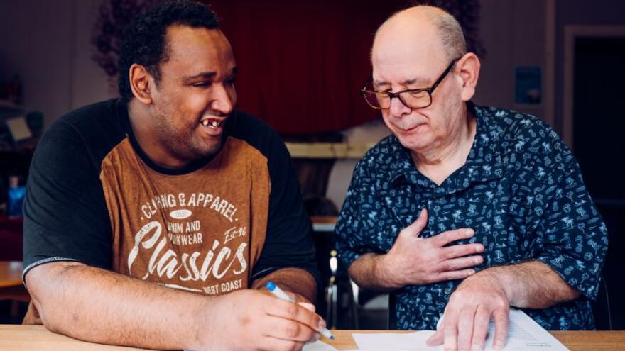 Two men sitting at a desk talking and writing in a note pad