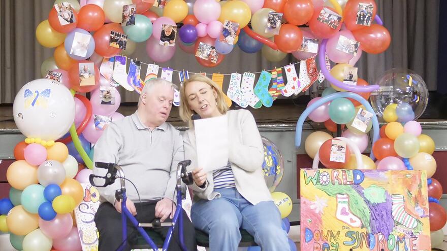 man and woman with a microphone in front of lots of multicoloured balloons at a party
