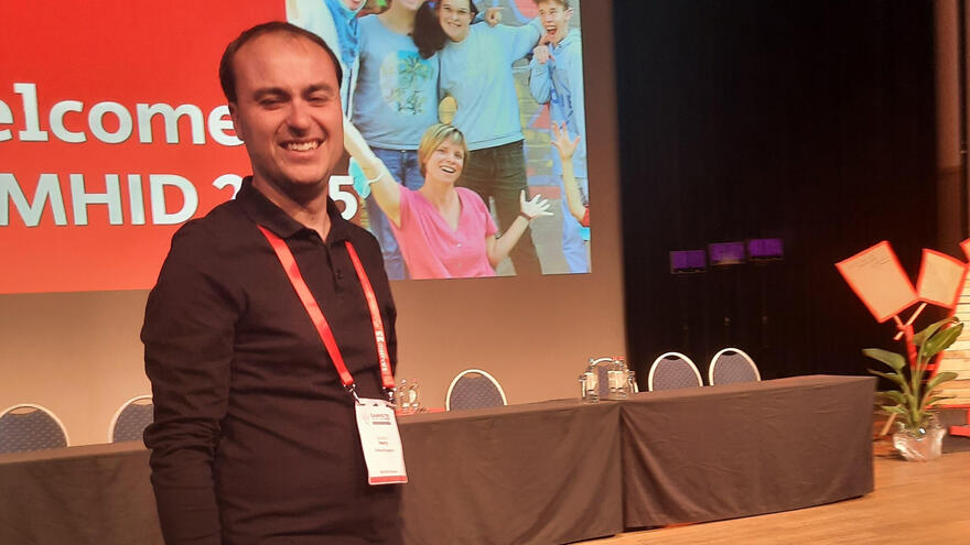 A man in a black top onstage smiling, with a large screen behind him.