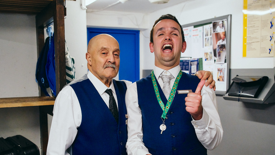A picture of two men standing next to each other. One man is laughing and has his thumbs up.