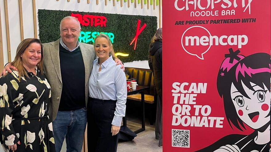 3 people standing in a Chopstix restaurant next to a Mencap and Chopstix banner
