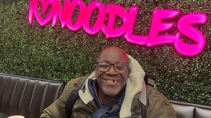 A man with a learning disability sitting in a Chopstix restaurant underneath a sign which says I love noodles
