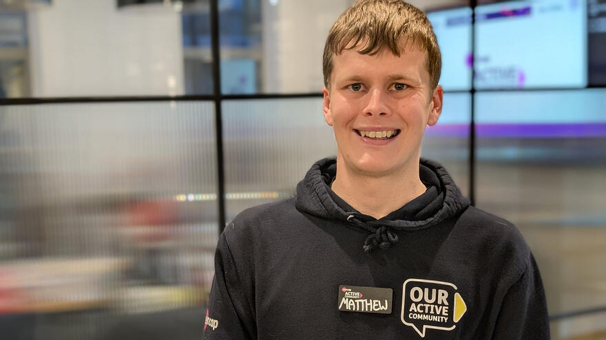 Matthew has short hair and is smiling widely. He is standing in front of a glass wall and is wearing a black hoody with the Mencap logo on the sleeve and Our Active Community on the chest