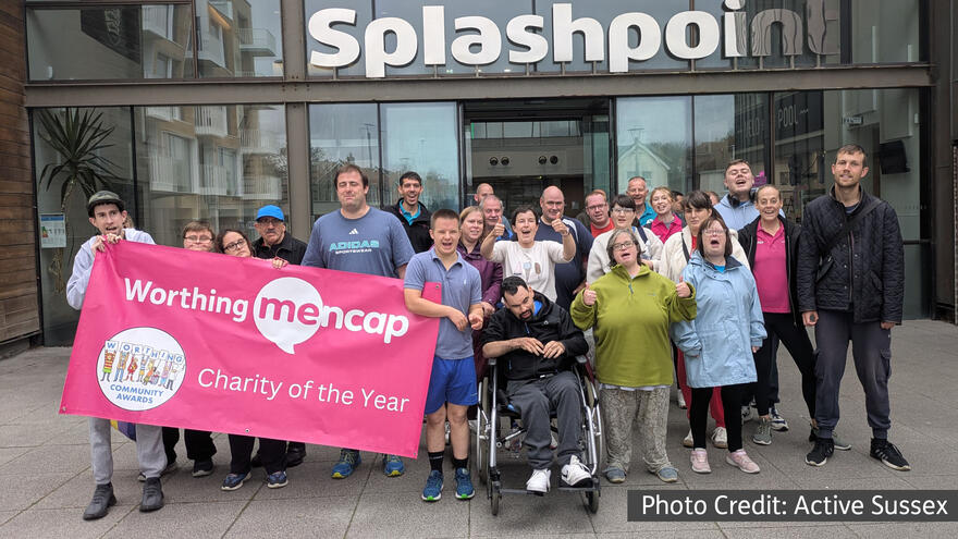 A group of people standing outside a leisure centre. A few on the left are holding a pink banner with the Worthing Mencap logo on it