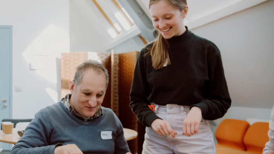A man at a table working on a painting. Next to him is a woman standing up and smiling at him