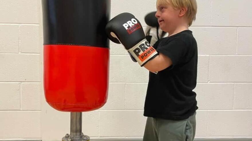 A little boy in a black t-shirt with boxing gloves on in front of a standing punch bag