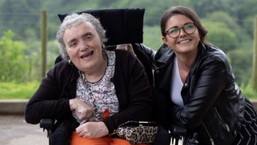 An older lady in a wheelchair smiles, next to her in a younger woman wearing glasses
