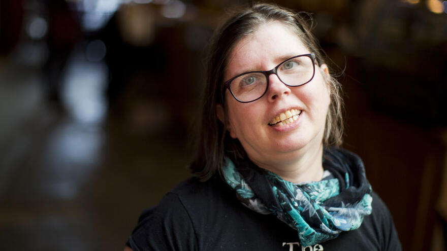 Amy has shoulder length brown hair and glasses. She is smiling at the camera nd wearing a black t shirt and green scarf.