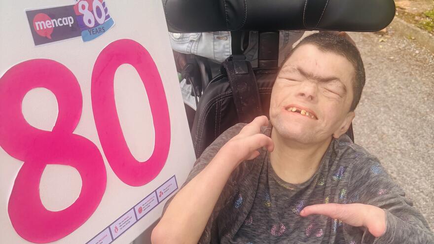 A man in a wheelchair and a carer behind him are outside holding a 'Mencap 80' sign