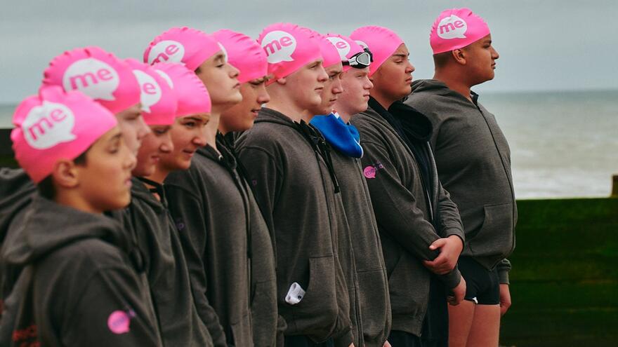 swimmers in Mencap swimming hats standing in a line on a beach