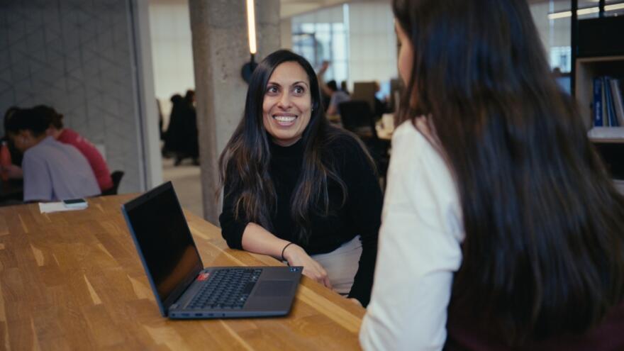 Two women sitting at a desk in front of a laptop talking to each other