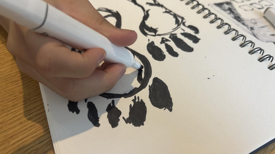 A hand holding a large artist pen is drawing an outline of feet onto paper