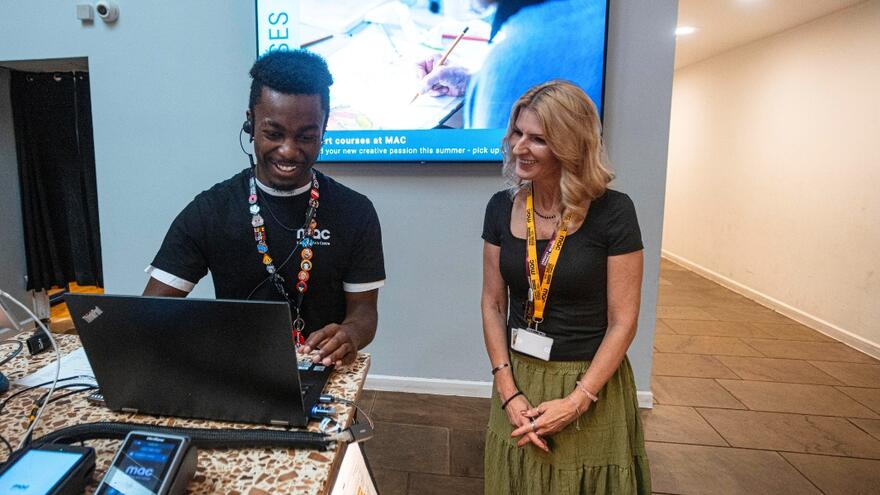 A young man wearing a black t-shirt and lanyard is working at a till counter with a female colleague next to him