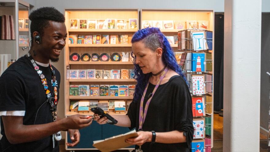 A young man wearing a black t-shirt and lanyard is working with a colleague in a gift shop part of a theatre