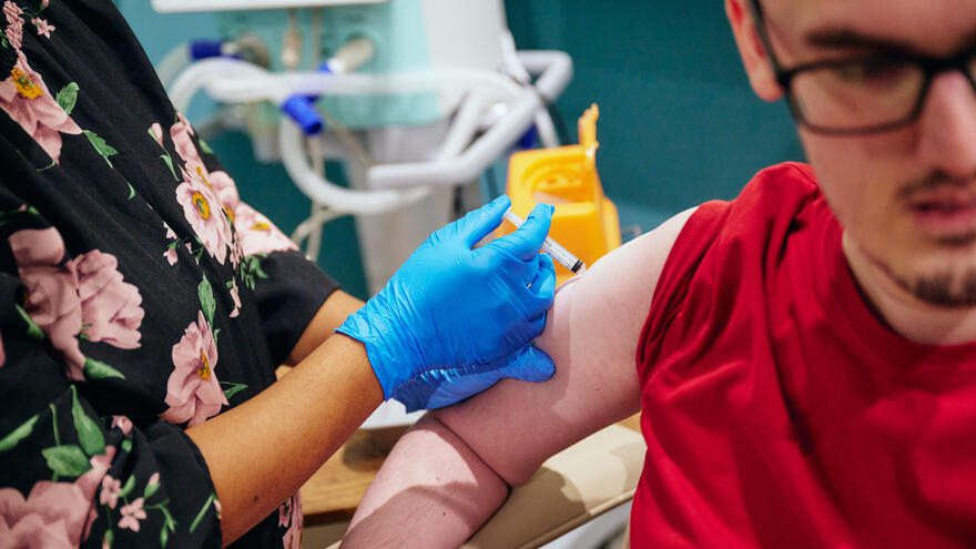 A close up of a nurses hand wearing blue clinical gloves giving a flu jab vaccination into the arm of a young man wearing glasses and a red top