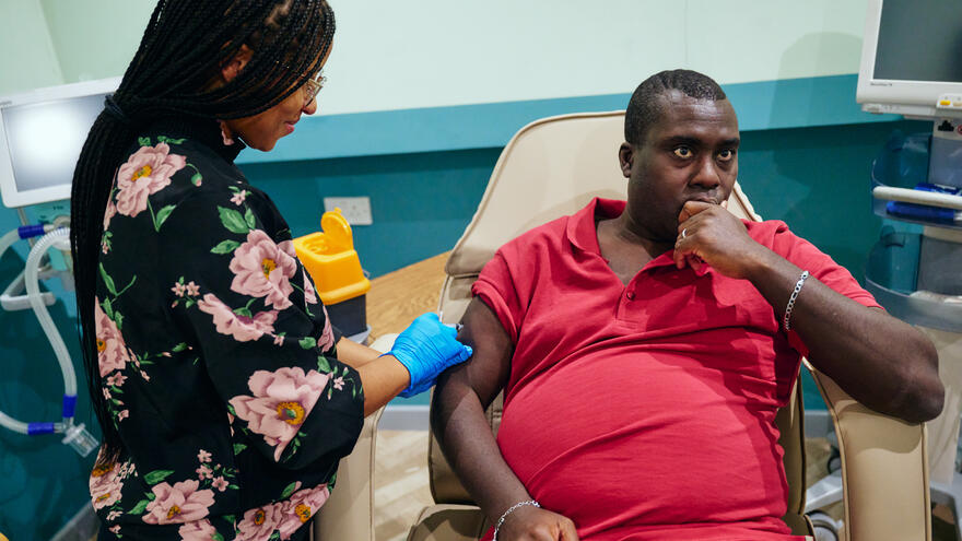 A nurse stands wearing blue clinical gloves and is giving an flu jab vaccination into the arm of a man sitting in a chair wearing a red top