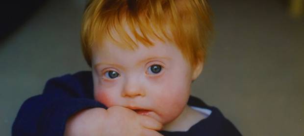 Baby boy with ginger hair wearing a blue jumper looking into camera with his hand in his mouth.