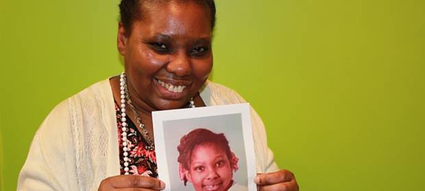 Woman stood against green background smiling and holding a photo of herself as a young girl.