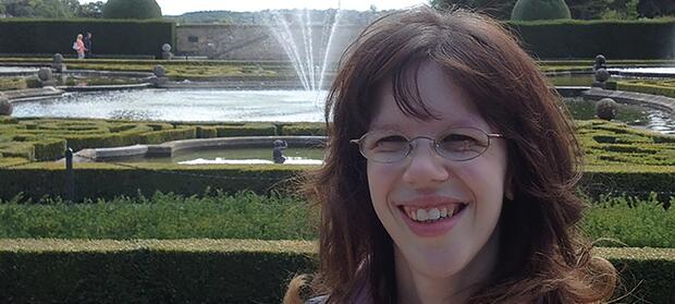 Woman with brown hair and glasses stood in park gardens in front of a water fountain, smiling into camera.