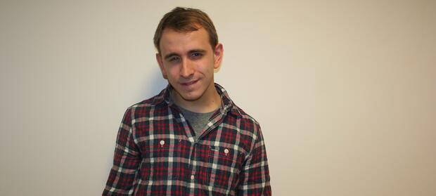 Young man wearing  chequered shirt stood in front of white wall.