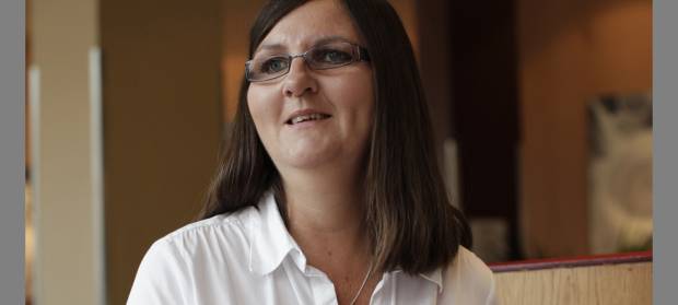 Woman with long brown hair and glasses, wearing a white shirt smiling and looking into the distance.
