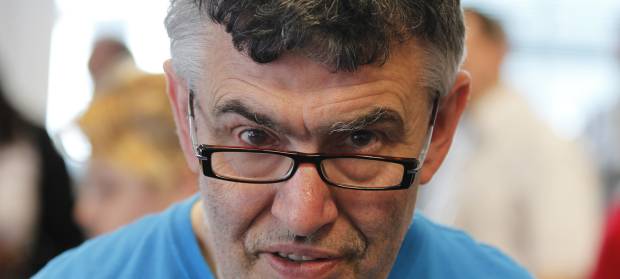 Man with grey curly hair and glasses, wearing a blue top, looks at camera. 