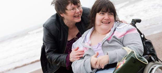 Two women smiling together on a beach, one woman is using a wheelchair whilst the other stands next to her, with the ocean in background