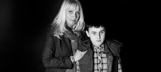 Woman stood with her hands on her son's shoulders who is stood in front of her.