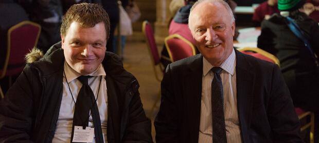 Two men wearing black suits sat at table next to each other smiling for camera.