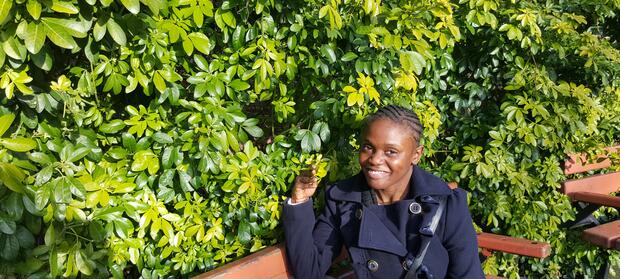 young woman sat on bench outside in front of bush. She is wearing a black coat and is looking up and smiling into the camera.
