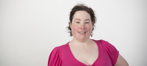 Woman with short curly dark hair wearing pink top stood in front of white background smiling into camera.