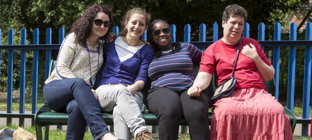 Four women sat together outside in the sunshine on a park on a bench, blue metal fence railing in the background