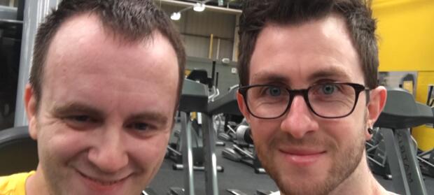 Two smiling young men stood next to each other in gym.