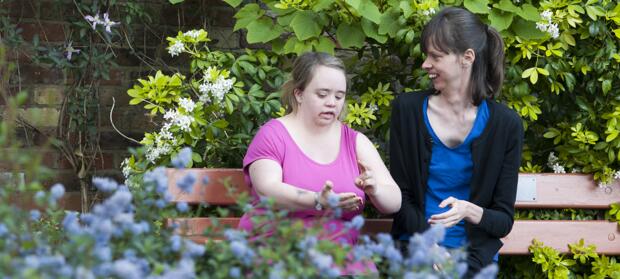 Two women sat together on bench in front of green bushes.
