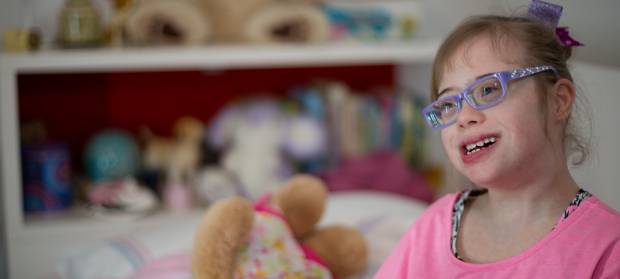Young girl with glasses wearing a pink top sat in bedroom with toys around her, smiling.