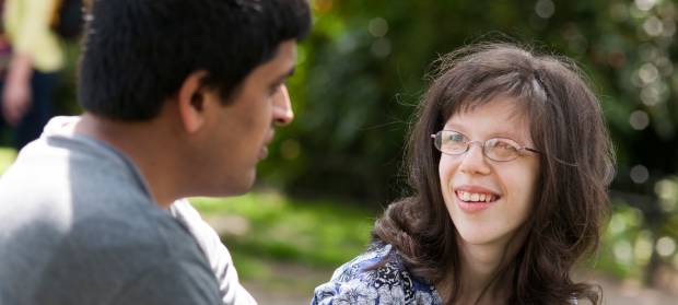 Man and woman sat outside together in park. The woman has long dark hair and glasses and is smiling at the man next to her.