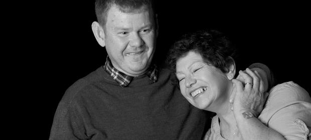 Man and woman stood together smiling. The man has his arm around the woman's shoulder.
