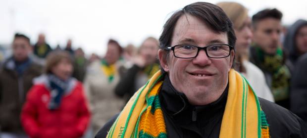 Man with glasses stood in crowd outside at football match, wearing a yellow and green football scarf, smiling at the camera