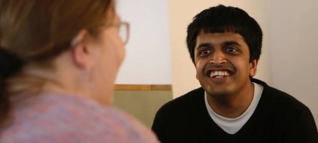 Young man with dark hair wearing black jumper smiling looking at person sat opposite him.