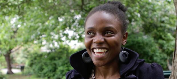 Young woman stood outside wearing dark coat and large earrings, smiling.
