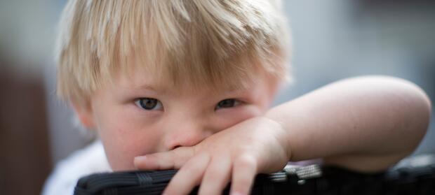 Young boy with blond hair leaning his face on back of chair with his arm in front of his face