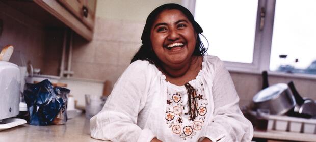 Woman with dark hair wearing white top stood by kitchen counter, laughing.