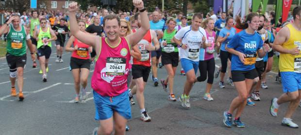 Man running wearing a Mencap vest raising his arms in the air. Lots of other runners in the background.