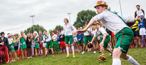 Team of people stood in field playing dodgeball, crowds of people in the background watching.