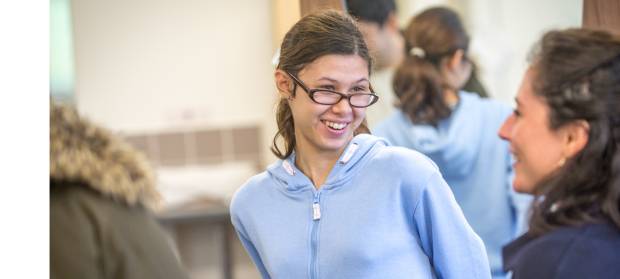 Young woman with hair tied in a ponytail wearing glasses and a pale blue jumper smiling.