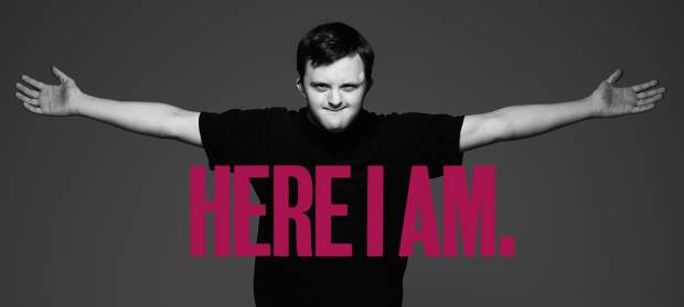 Young man wearing black t-shirt stood holding his arms out wide. Pink text reading "Here I am" in front of him.