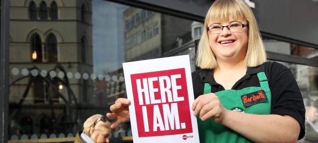 Woman wearing Starbucks uniform, stood outside coffee shop holding a Here I am sign.