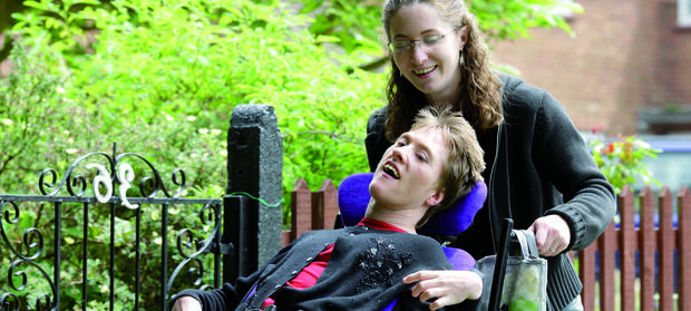 Two women outside. One woman is in a wheelchair which is being pushed by the other woman behind her.