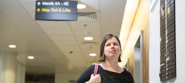 Woman walking down hospital corridor holding bag on her shoulder.