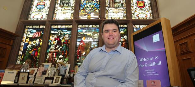 Man wearing pale blue shirt sat in front of stained glass window.